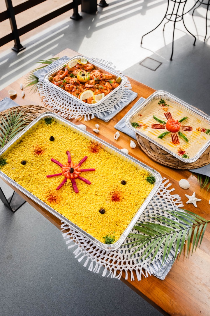 A table set with three large catering trays filled with vibrant dishes. The centerpiece is a tray of yellow rice garnished with vegetables arranged in a decorative star pattern. Surrounding it are trays of shrimp and a creamy dish, all beautifully displayed with tropical leaves and seashell decorations.