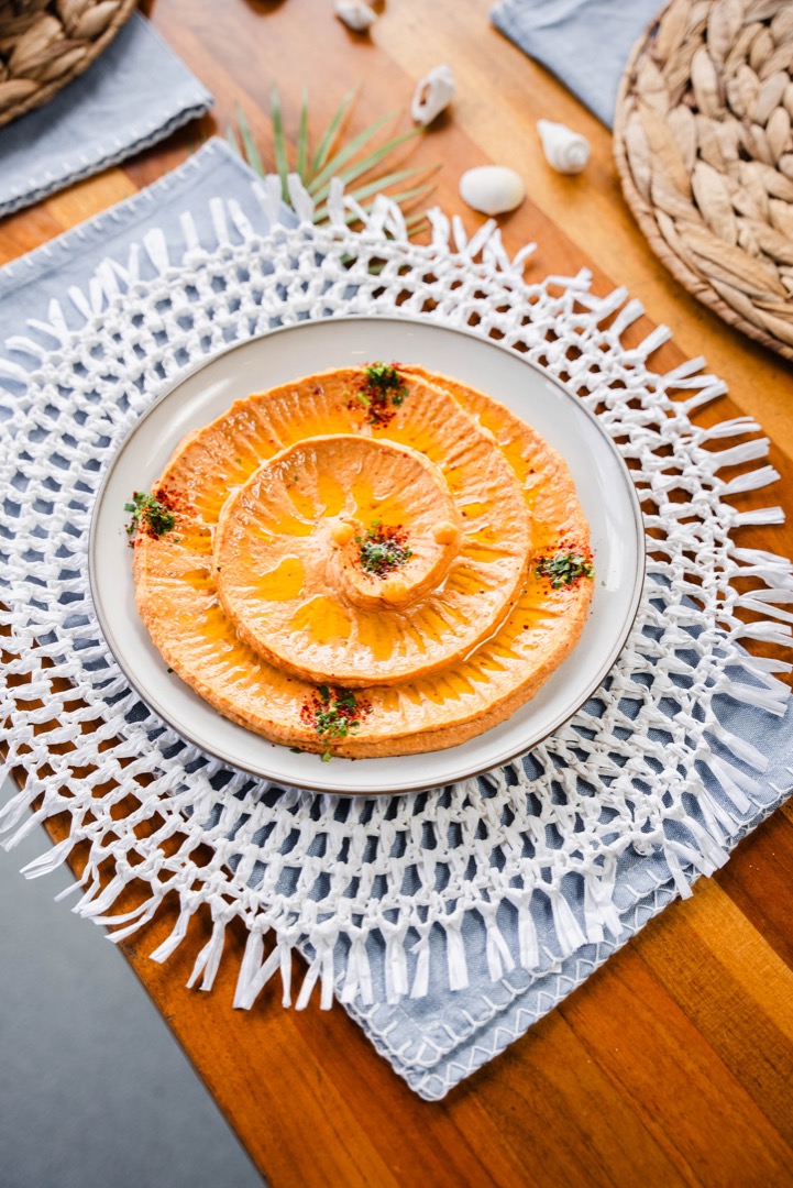 A beautifully plated dish of thinly sliced oranges arranged in a concentric pattern, garnished with herbs and a light dressing, presented on a neutral-colored plate on top of a woven placemat with coastal decor elements.