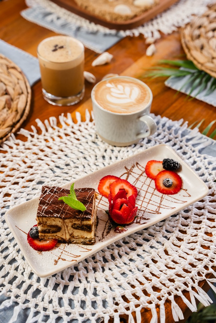 A plate of tiramisu dessert garnished with sliced strawberries and a sprig of mint, elegantly plated with a drizzle of chocolate. Accompanying the dessert are two coffee beverages: one cup with latte art and another glass with iced coffee. The table setting is styled with a woven placemat and natural décor, creating a cozy café ambiance.