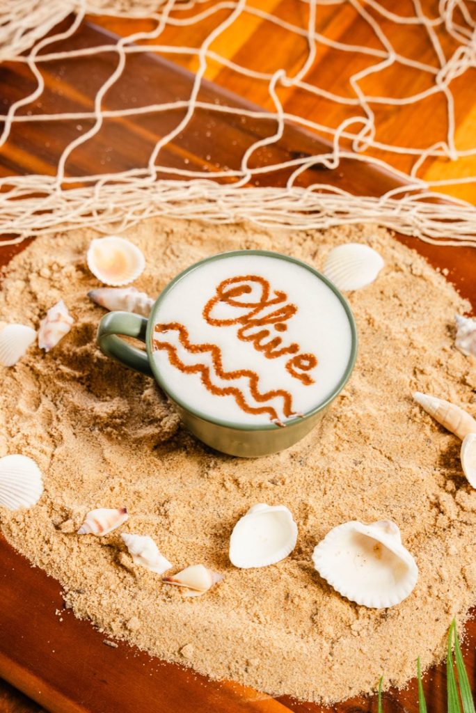 A cup of coffee with latte art that spells "Olive" placed on a bed of sand, surrounded by seashells and decorative fishing net, creating a beach-themed aesthetic.