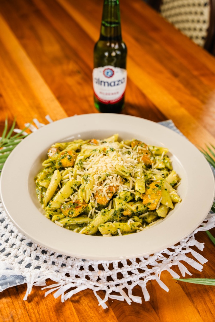 A plate of pesto penne pasta garnished with shredded cheese, served on a white dish. The pasta is mixed with grilled vegetables, creating a vibrant, appetizing dish. Behind the plate is a bottle of Almaza Pilsner beer, set on a wooden table, adding a rustic touch to the presentation. The overall scene captures a relaxed, inviting dining atmosphere.