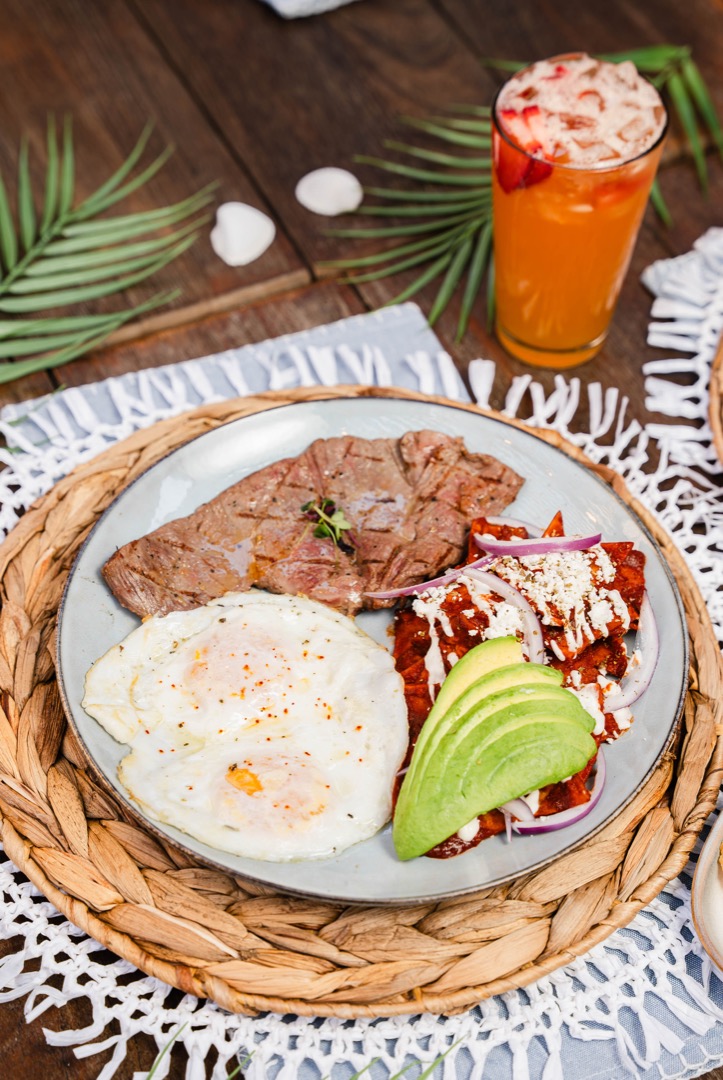 A breakfast plate featuring steak, fried eggs, avocado slices, and chilaquiles topped with cheese, served on a woven placemat.