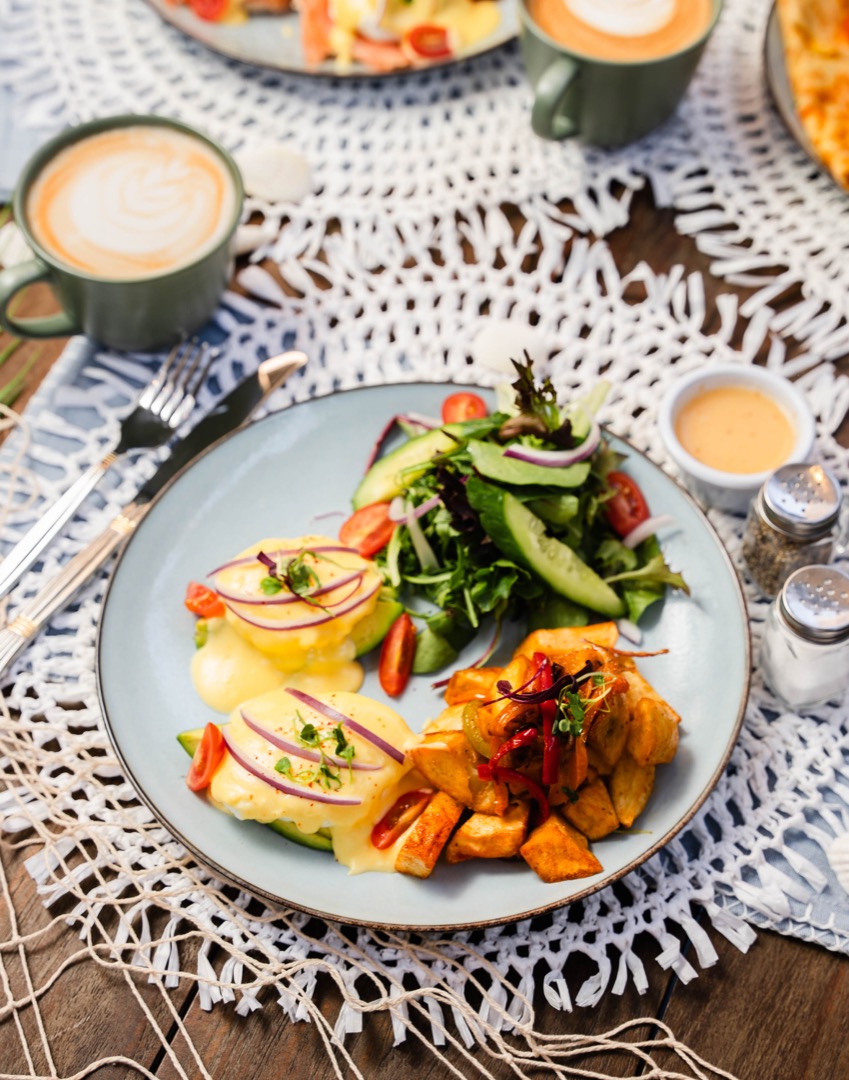 A breakfast plate featuring eggs benedict topped with hollandaise sauce and garnished with microgreens, accompanied by a fresh garden salad with avocado slices and a side of roasted potatoes. The dish is served on a light blue plate placed on a woven white placemat with a rustic wooden table in the background. Two lattes are seen in green mugs nearby, completing the cozy dining setup.