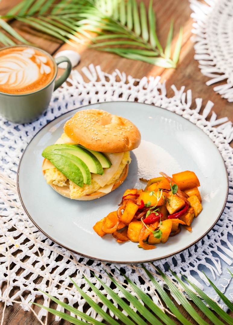 A breakfast plate featuring a bagel sandwich layered with egg, avocado slices, and melted cheese, served with a side of roasted breakfast potatoes. The plate is presented on a woven white placemat with tropical palm leaves in the background. A beautifully crafted latte in a green mug complements the meal, adding a warm touch to this inviting brunch scene.