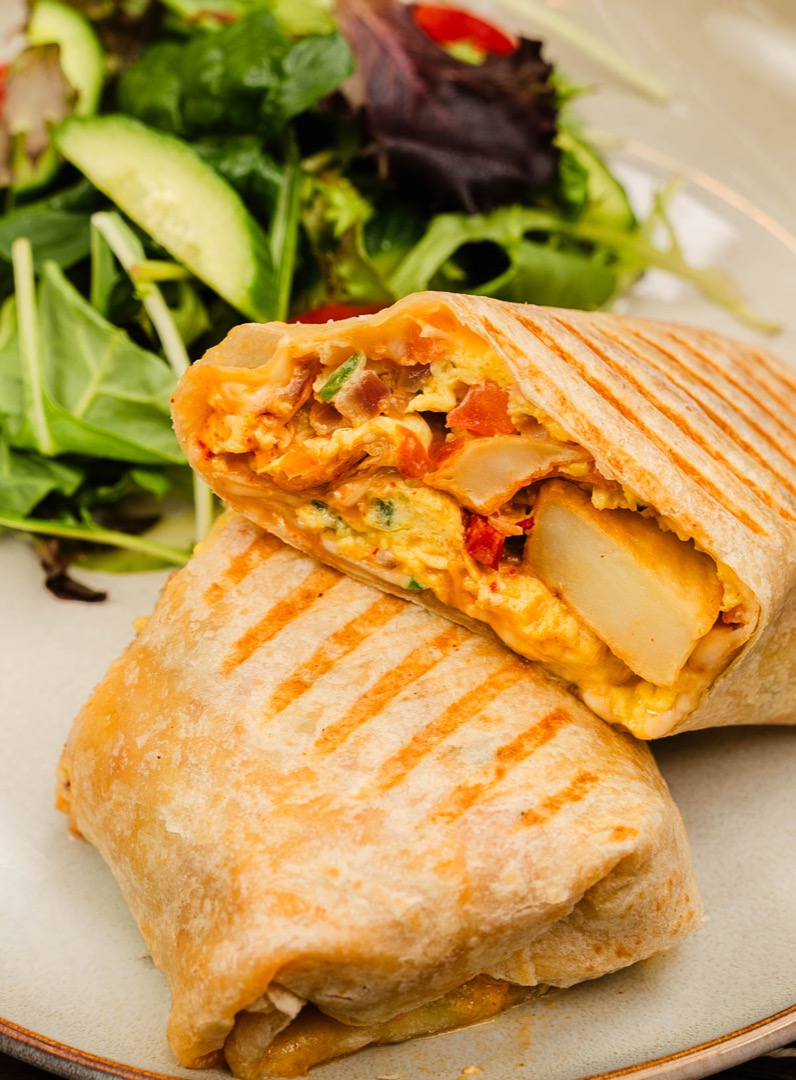 A close-up of a grilled breakfast burrito cut in half, revealing a filling of eggs, potatoes, cheese, and vegetables, with a side salad in the background.