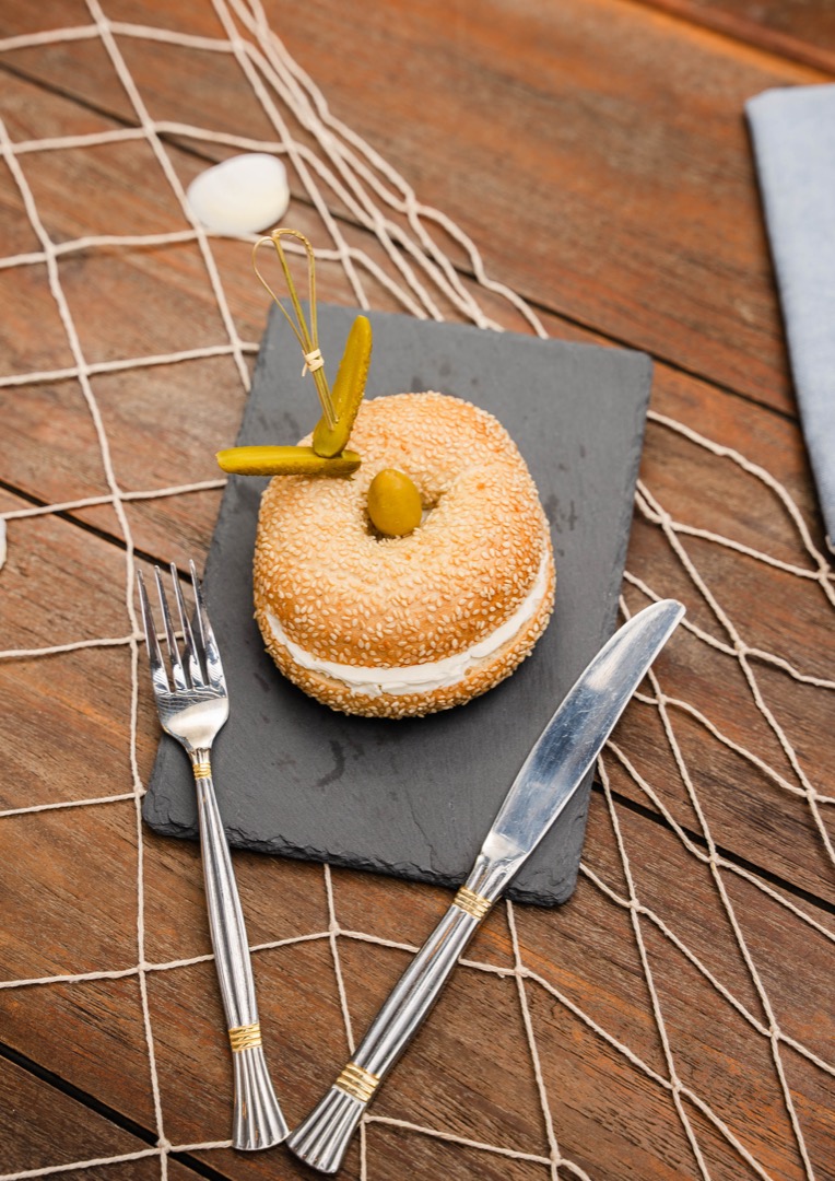 A small sandwich on a sesame seed bun, garnished with an olive and a pickled pepper, placed on a slate serving board with a fork and knife.