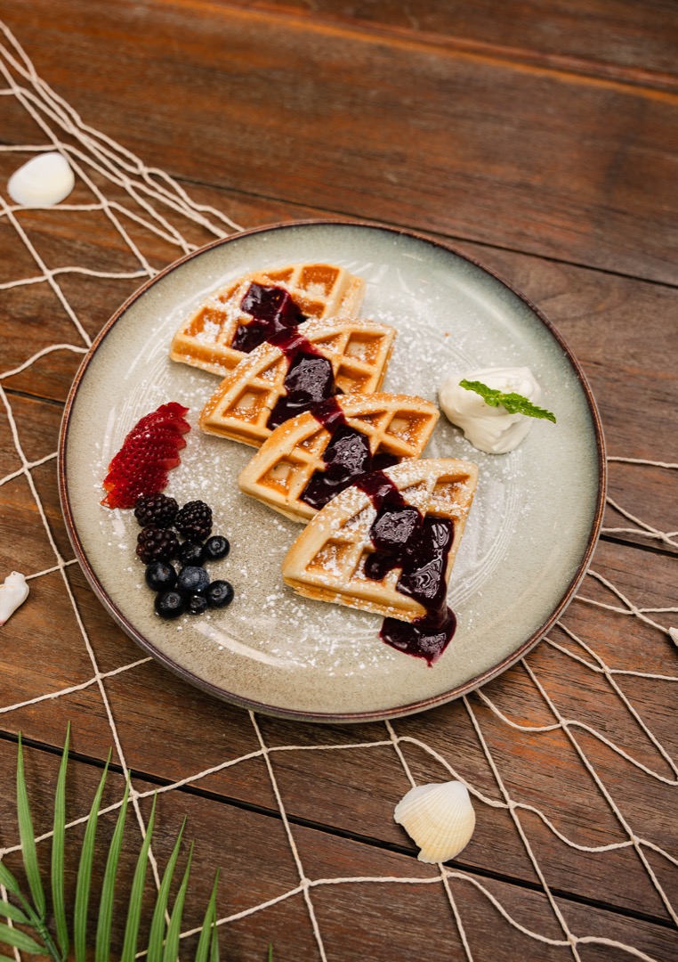 A plate of waffles topped with berry compote and powdered sugar, served with fresh berries and whipped cream.