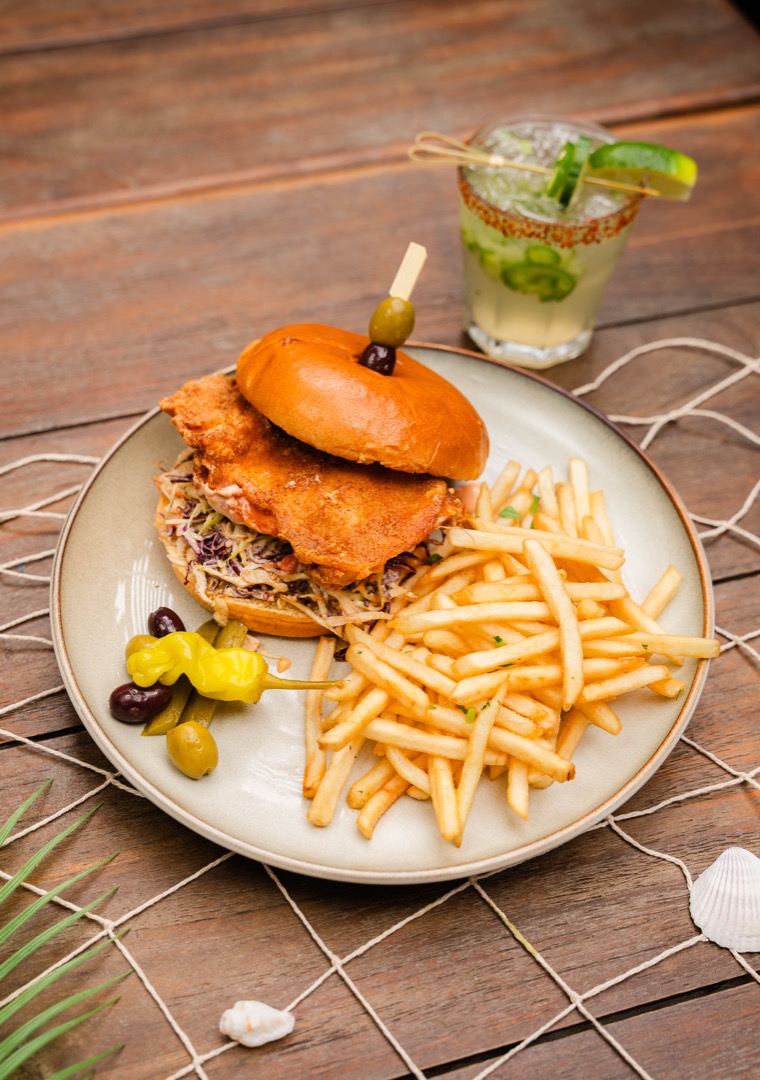 A fried chicken sandwich served on a brioche bun with coleslaw, accompanied by a side of crispy fries and pickled peppers, with a refreshing cocktail garnished with cucumber and lime in the background.