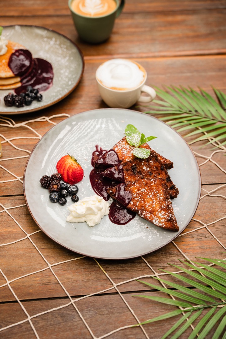 A plate with French toast topped with berry compote, powdered sugar, and mint leaves, served with a side of whipped cream and fresh berries.