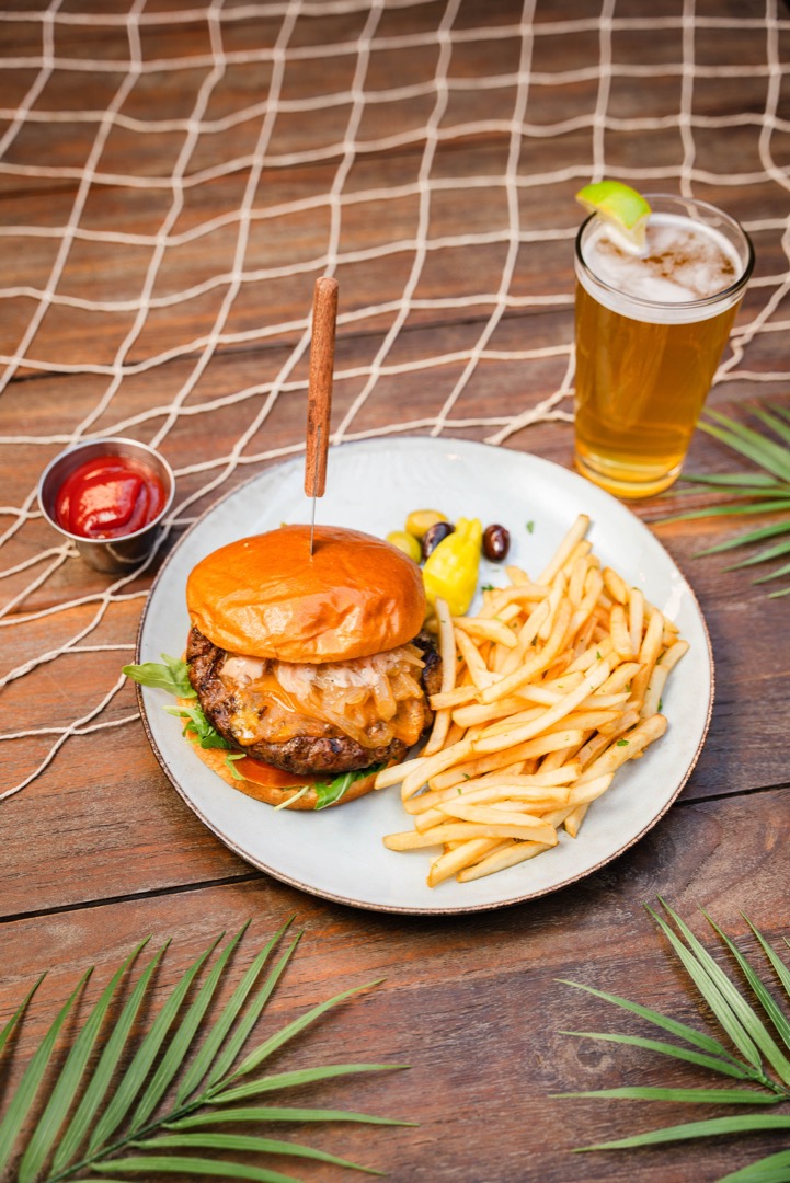 A close-up view of a gourmet burger served at Olive by the Bay. The burger features a juicy patty topped with caramelized onions and cheese, served on a toasted bun with fresh greens. Accompanying the burger is a generous serving of crispy French fries, olives, a pepperoncini, and a side of ketchup. A tall glass of beer with a lime wedge sits beside the plate. The meal is presented on a white plate set on a wooden table decorated with tropical palm leaves and a fishing net, enhancing the coastal dining ambiance.