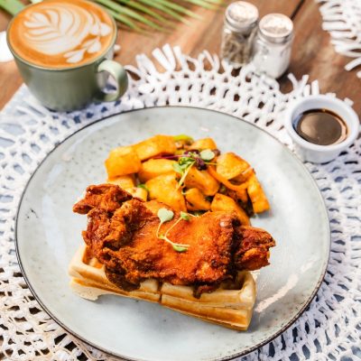 A plated dish of crispy fried chicken served on top of golden waffles, accompanied by seasoned roasted potatoes. The meal is set on a woven white placemat with a green mug filled with a frothy latte featuring foam art in the background. A small cup of syrup is placed next to the plate, with tropical leaves adding a fresh vibe to the rustic table setting.