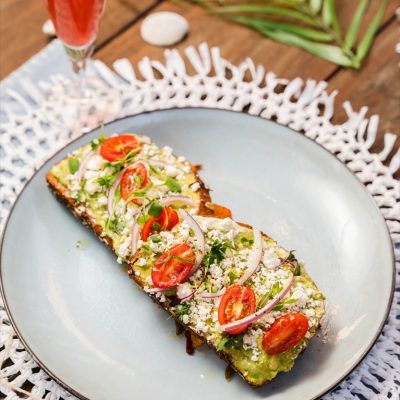 A plate featuring a slice of avocado toast topped with sliced cherry tomatoes, red onions, crumbled feta cheese, and fresh herbs. The dish is accompanied by a vibrant red-orange beverage served in a champagne flute, creating a visually appealing brunch setup. The plate rests on a woven placemat with tropical palm leaves in the background, enhancing the coastal brunch ambiance.