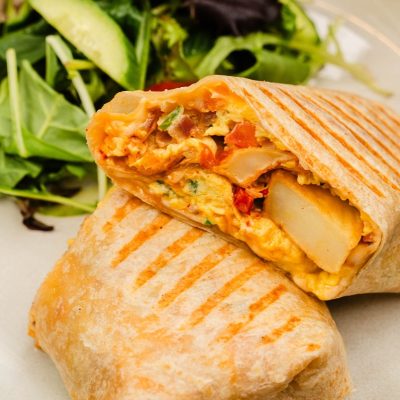 A close-up of a grilled breakfast burrito cut in half, revealing a filling of eggs, potatoes, cheese, and vegetables, with a side salad in the background.