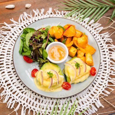 A plate of eggs benedict with avocado, served with roasted potatoes, a side salad, and a small dish of hollandaise sauce.