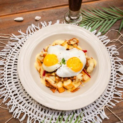 A breakfast plate featuring hash topped with two sunny-side-up eggs, served on a rustic table with a decorative crochet placemat and a glass of iced coffee.