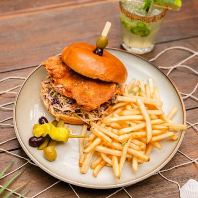 A fried chicken sandwich served on a brioche bun with coleslaw, accompanied by a side of crispy fries and pickled peppers, with a refreshing cocktail garnished with cucumber and lime in the background.