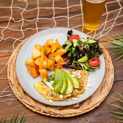 A breakfast plate featuring an avocado toast with feta cheese, served with crispy breakfast potatoes and a side salad of mixed greens, cherry tomatoes, and cucumbers, paired with a glass of beer with a lime wedge.