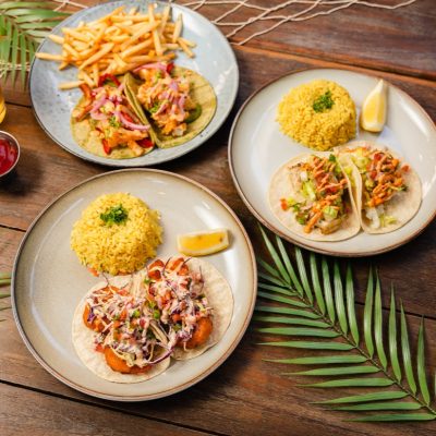 An overhead view of a taco trio and sides served at Olive by the Bay. The spread includes three plates of tacos: one with shrimp tacos topped with red onions and a side of French fries, another with fish tacos topped with coleslaw and served with yellow rice and a lemon wedge, and a third plate with two tacos filled with grilled chicken, lettuce, and sauce accompanied by yellow rice and a lemon wedge. The table is decorated with tropical palm leaves and a fishing net, enhancing the coastal dining experience.