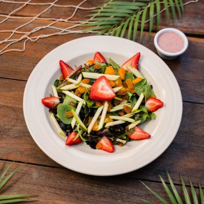 A close-up view of a fresh and vibrant salad served at Olive by the Bay. The dish features mixed greens topped with sliced strawberries, julienned apple slices, golden raisins, and orange segments. A small bowl of dressing is placed next to the plate. The salad is set on a wooden table decorated with tropical palm leaves and a decorative fishing net, enhancing the coastal dining ambiance. The colorful ingredients highlight the freshness and healthiness of the dish.