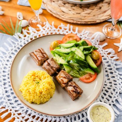 A delicious Mediterranean-style dish featuring grilled beef skewers, fluffy yellow rice, and a fresh cucumber and tomato salad on a light ceramic plate.