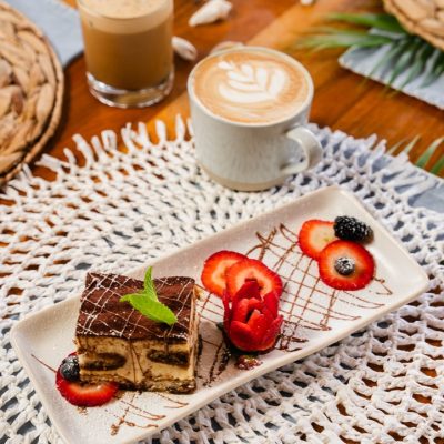 A plate of tiramisu dessert garnished with sliced strawberries and a sprig of mint, elegantly plated with a drizzle of chocolate. Accompanying the dessert are two coffee beverages: one cup with latte art and another glass with iced coffee. The table setting is styled with a woven placemat and natural décor, creating a cozy café ambiance.