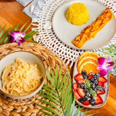 An overhead view of a vibrant table setting featuring a variety of dishes. The spread includes a plate with saffron rice and grilled salmon, a bowl of creamy pasta, and a colorful fruit bowl with sliced oranges, strawberries, and blackberries, garnished with a purple orchid. The dishes are set on decorative placemats and surrounded by tropical leaves, creating a refreshing and inviting ambiance.