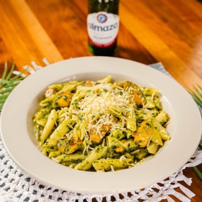 A plate of pesto penne pasta garnished with shredded cheese, served on a white dish. The pasta is mixed with grilled vegetables, creating a vibrant, appetizing dish. Behind the plate is a bottle of Almaza Pilsner beer, set on a wooden table, adding a rustic touch to the presentation. The overall scene captures a relaxed, inviting dining atmosphere.